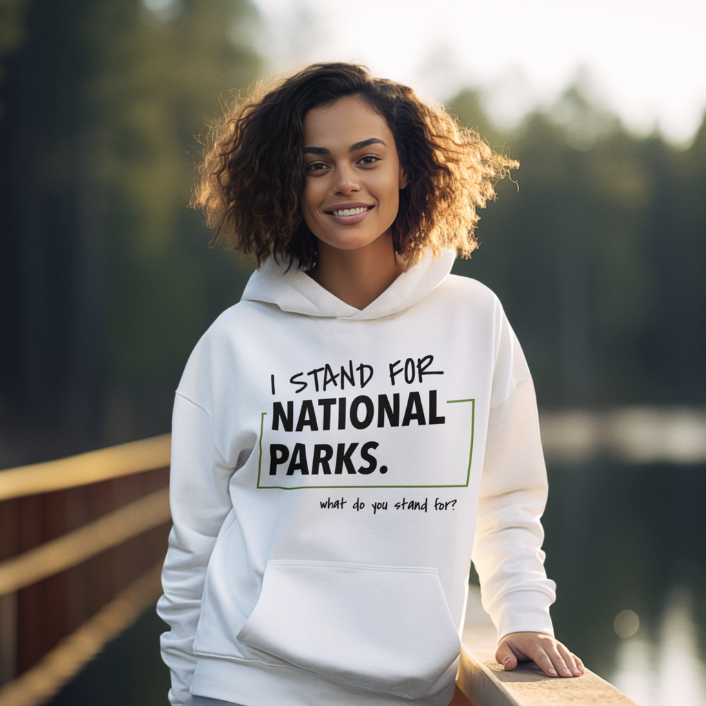 Woman wearing a white hoodie with 'I stand for National Parks' text, standing by a lake.
