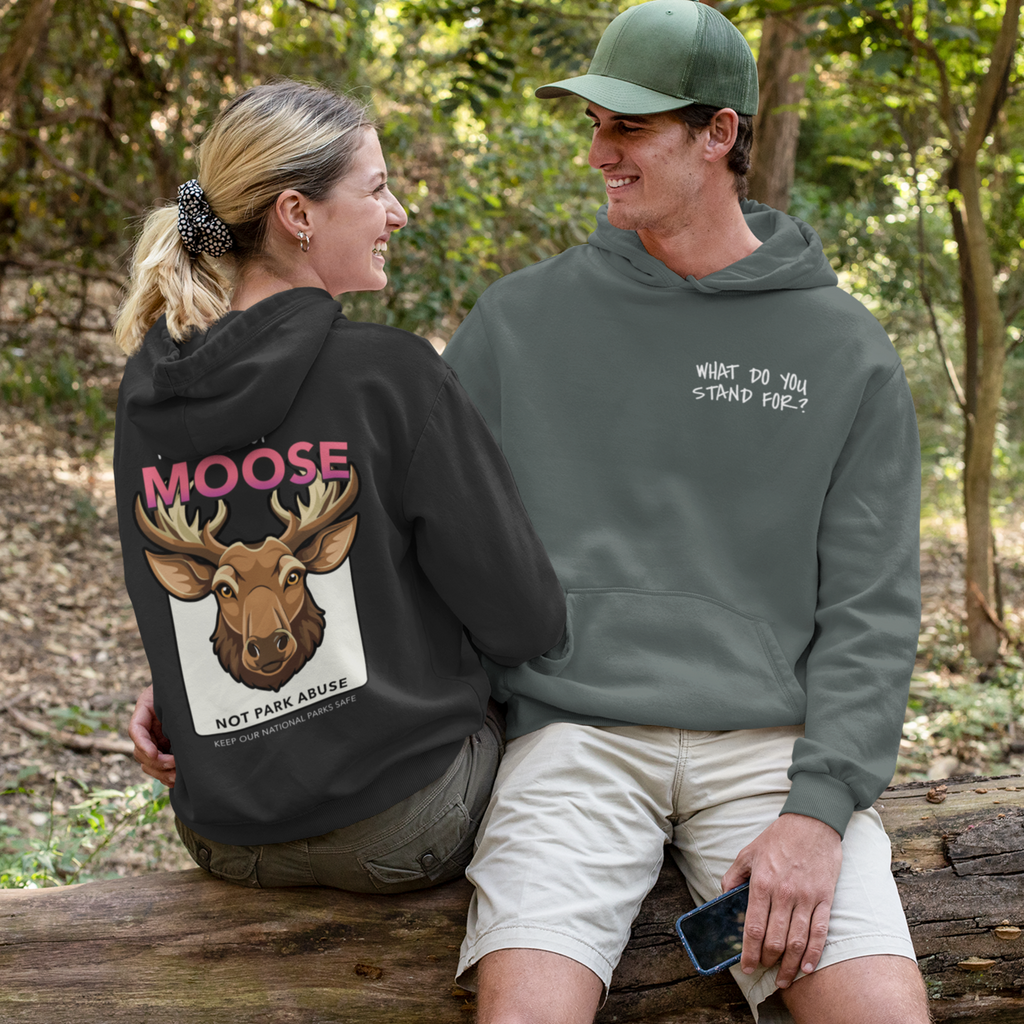 Two people sitting on a log outdoors, one wearing a black hoodie with a moose graphic and text, the other wearing a green hoodie with text.