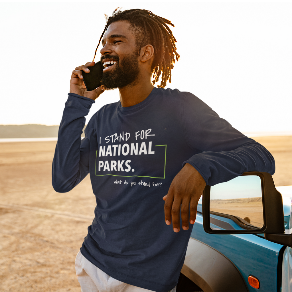 Man in a 'I Stand for National Parks' shirt talking on a phone by a car in a desert setting