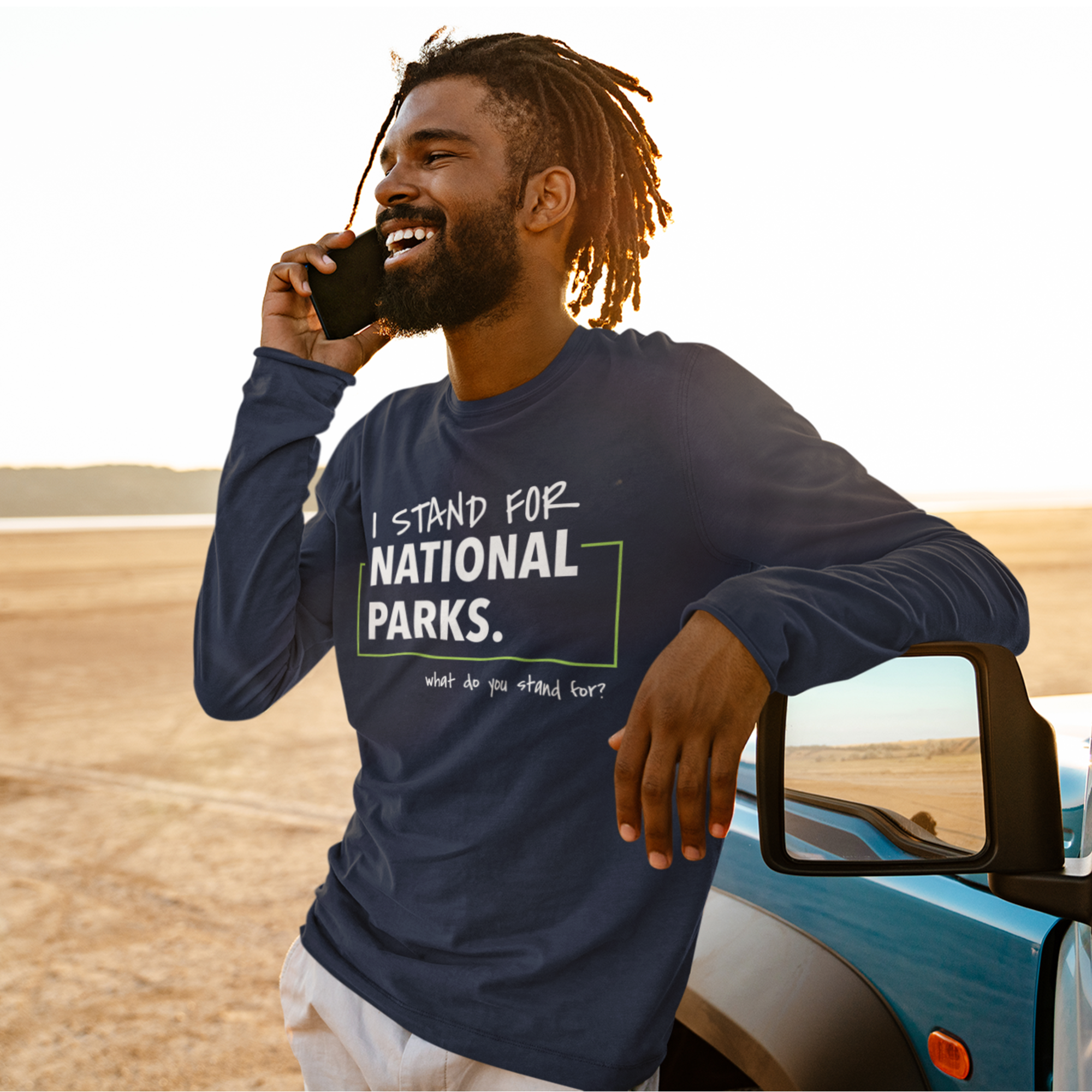Man in a 'I Stand for National Parks' shirt talking on a phone by a car in a desert setting