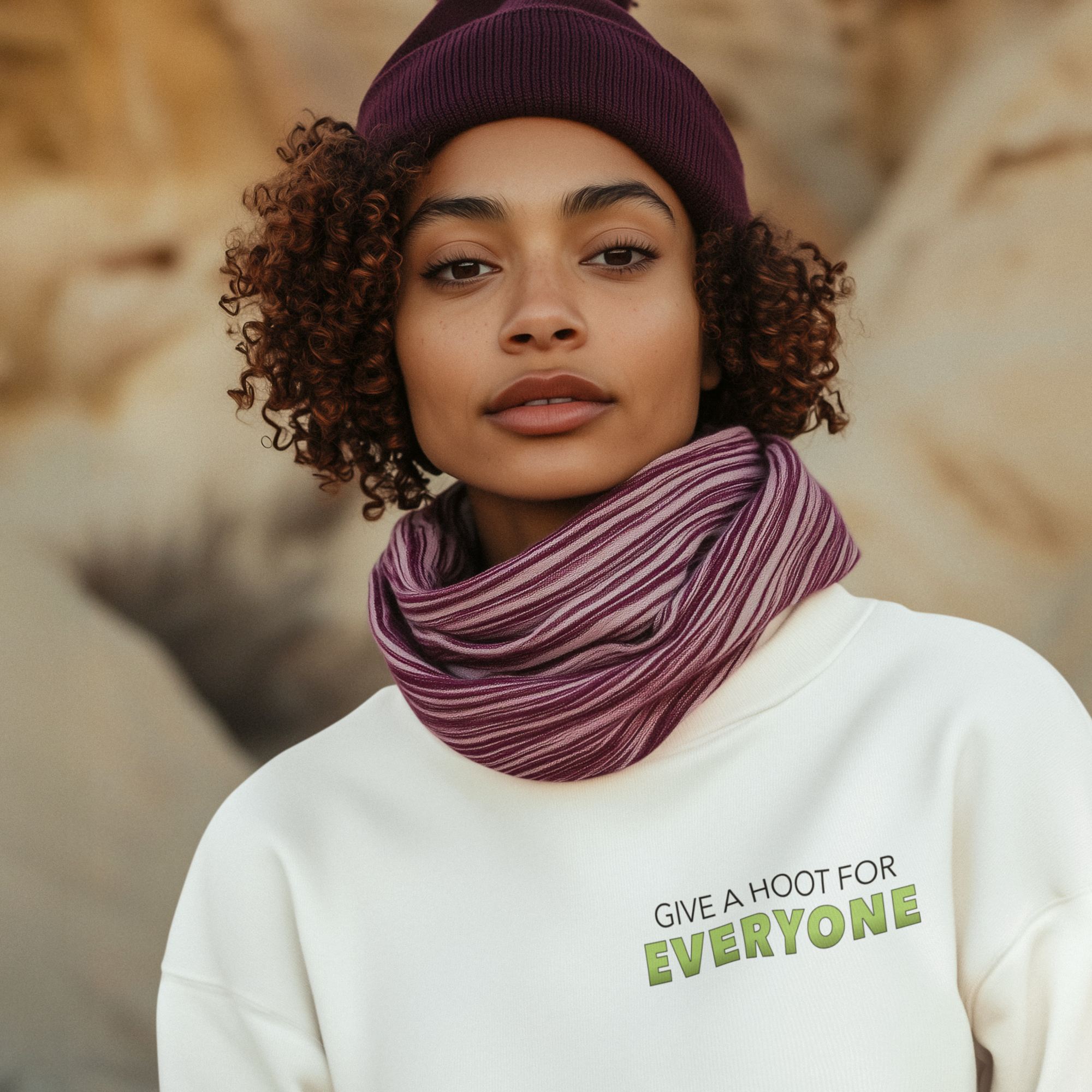 Person wearing a white sweatshirt with 'Give a Hoot for Everyone' text, purple beanie, and scarf against a natural background.
