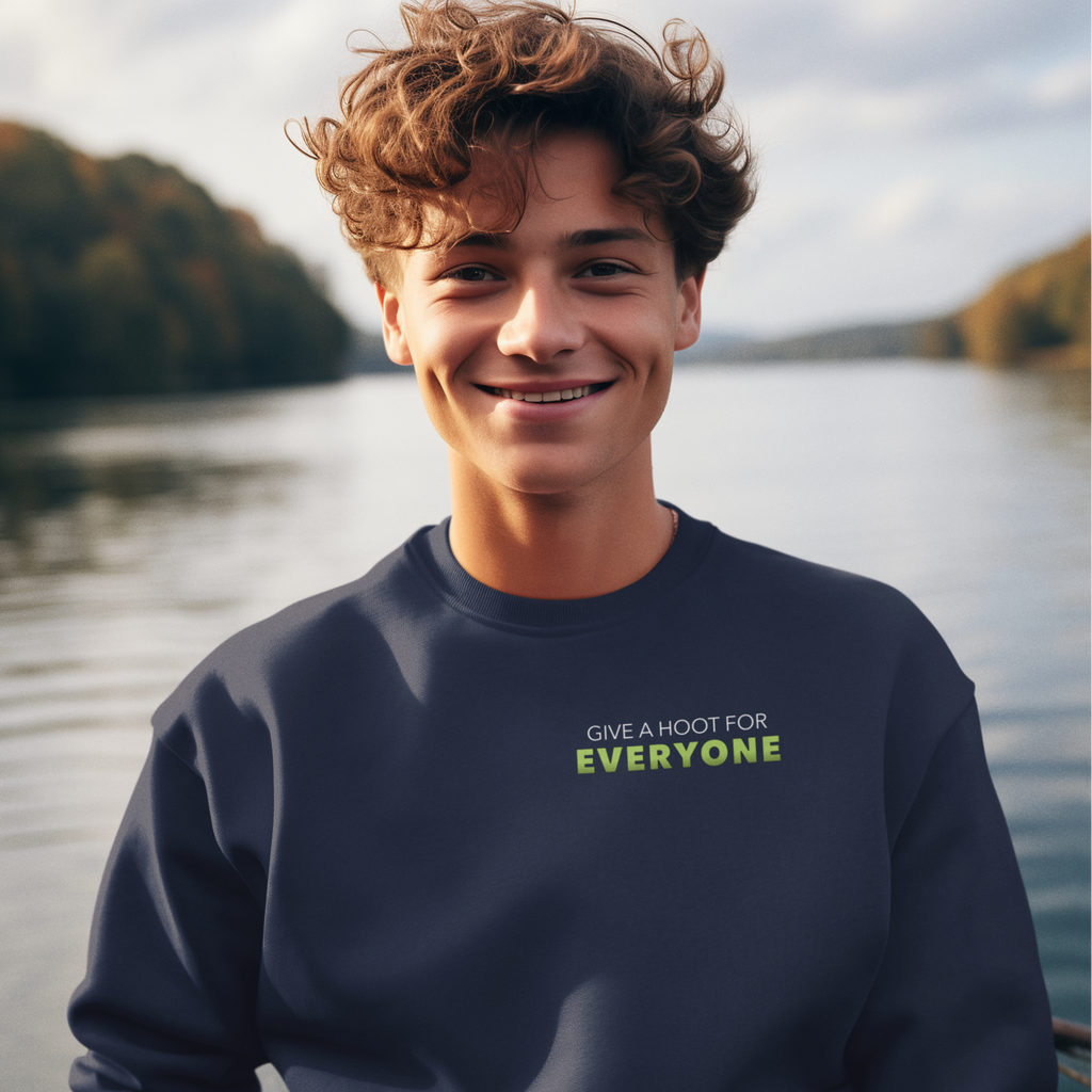 Person wearing a navy sweatshirt with 'Give a Hoot for Everyone' text by a lake.
